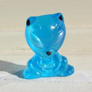 May include: A translucent blue alien figurine with iridescent eyes and black accents. The figure has a bear-like shape with large eyes and small ears. The figurine is sitting down, with its arms resting on its legs.