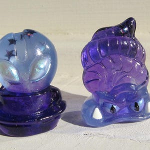 Puede incluir: Dos figuras translúcidas de resina. Una es un alien azul con detalles de estrellas, la otra es una criatura morada con una cabeza bulbosa y detalles blancos. Ambas tienen un acabado brillante y están sobre una superficie blanca.