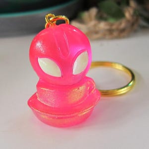 May include: A vibrant pink keychain featuring a small alien figure with white eyes. The alien is perched on a saucer-shaped base, with a gold-colored ring attached. The keychain has a metallic sheen.