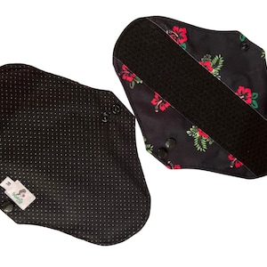 Op de afbeelding: Twee zwarte herbruikbare stoffen pads met een bloemenprint. Eén pad heeft een zwarte klittenbandstrip en de andere heeft een zwart stippenpatroon.
