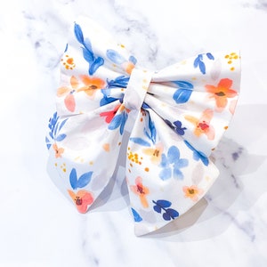 May include: A white fabric bow with a floral pattern in shades of blue, orange, and pink. The bow is tied in a classic style with a center loop.