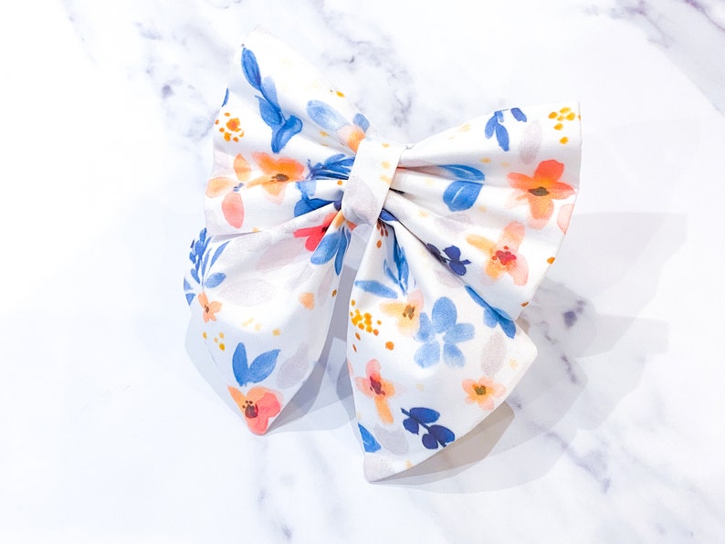 May include: A white fabric bow with a floral pattern in shades of blue, orange, and pink. The bow is tied in a classic style with a center loop.