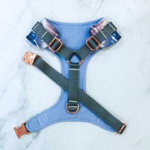 May include: A light blue dog harness with a tie-dye pattern and rose gold hardware. The harness has a grey adjustable strap.