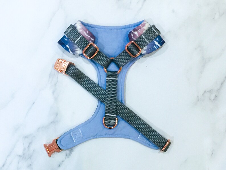 May include: A light blue dog harness with a tie-dye pattern and rose gold hardware. The harness has a grey adjustable strap.