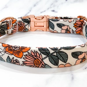 May include: A white dog collar with a floral pattern featuring orange, pink, and green flowers. The collar has a rose gold buckle and is made of durable fabric.