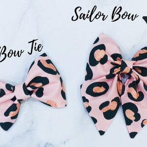 May include: Two pink and black leopard print fabric bows. The smaller bow is a bow tie and the larger bow is a sailor bow.