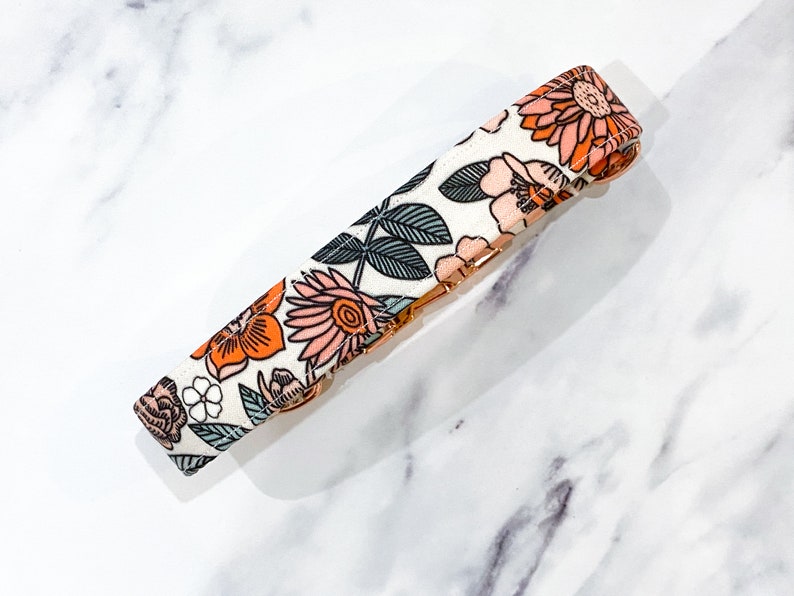 May include: A white dog collar with a floral pattern featuring orange, pink, and green flowers. The collar has a gold metal buckle.
