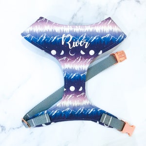 May include: A purple, blue, and white dog harness with a mountain and moon pattern. The harness has a rose gold buckle and the name "River" is printed on the front.