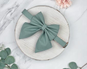 May include: A sage green bow tie with a matching adjustable strap and gold-colored hardware. The bow tie is centered on a white, rustic wooden plate, with floral and greenery accents.