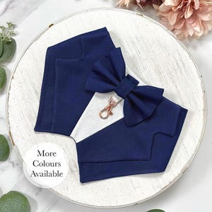May include: A navy blue fabric pet bandana designed to resemble a tuxedo jacket with a white triangular insert and a matching navy blue bow tie. A rose gold metal lobster clasp is attached to the center. The text More Colours Available is visible.