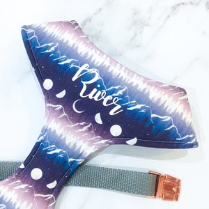 May include: A purple and blue dog harness with a mountain and moon pattern. The harness has the name "River" embroidered on it in white. The harness is attached to a gray leash with a rose gold buckle.