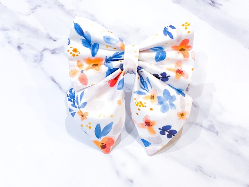 May include: A white fabric bow with a floral pattern in shades of blue, orange, and yellow. The bow is tied in a classic shape with a center loop.