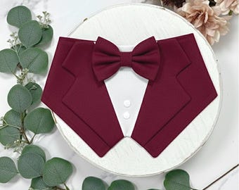 Puede incluir: Un pañuelo para mascota de color burdeos diseñado para parecer una chaqueta de esmoquin formal con un frente de camisa blanco, dos botones blancos pequeños y una pajarita burdeos a juego unida al cuello.