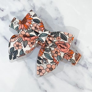 May include: A white bow tie with a floral pattern in shades of pink, orange, and green. The bow tie is made of fabric and has a gold metal clasp.