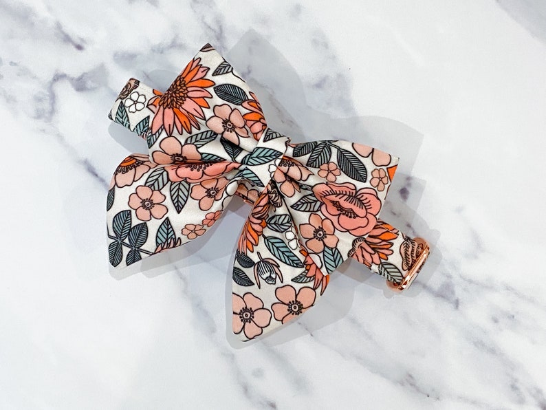 May include: A white bow tie with a floral pattern in shades of pink, orange, and green. The bow tie is made of fabric and has a gold metal clasp.