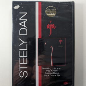 Könnte beinhalten: Eine schwarze DVD-Hülle für das Album "Aja" von Steely Dan. Das Cover zeigt den Albumtitel in einer stilisierten Schriftart mit roten und weißen vertikalen Streifen. Die DVD-Hülle ist in Kunststoff versiegelt.