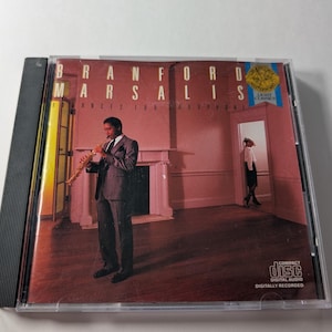 May include: A compact disc case featuring the album "Brantford Marsalis - Ances for Saxophone." The cover art depicts a man in a suit playing a saxophone in a room with a fireplace. The CD case is in a clear plastic case.