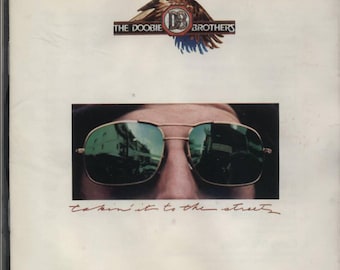 Puede incluir: Una portada de álbum vintage con un primer plano de una persona con gafas de sol. La portada del álbum es blanca con el texto "THE DOOBIE BROTHERS" y "Takin' It to the Streets" en rojo.