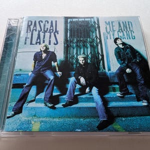 May include: A music CD case featuring the album cover for Rascal Flatts Me and My Gang. The cover shows three individuals sitting on blue concrete steps in front of a metal gate, wearing dark shirts and blue jeans, with the band and album title text.