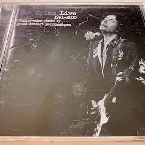 May include: CD case for Bob Dylan Live 1961-2000. The cover features a black and white image of Bob Dylan performing with a guitar and microphone. Text on the cover reads "Bob Dylan Live" and "Thirty-nine years of great concert performances."