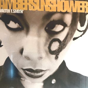 Può includere: Copertina di un album in bianco e nero con un primo piano di un volto con trucco occhi drammatico e rossetto scuro. Il testo "AMBERSUNSHOWER" e "WALTER T. SMITH" è stampato in oro.