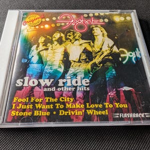 May include: A Foghat music CD titled Slow Ride and Other Hits in a clear plastic jewel case. The cover features a band performing on stage with yellow stage lights. Track titles listed include Fool For The City, I Just Want To Make Love To You, Stone Blue, and Drivin' Wheel, with the Flashback logo at the bottom.