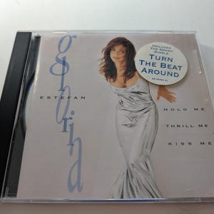 May include: A compact disc case featuring Gloria Estefan. The album cover shows a woman in a silver dress. The text on the cover includes "Gloria Estefan", "Hold Me", "Thrill Me", and "Kiss Me". A sticker reads "Turn the Beat Around".