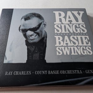 Puede incluir: Una portada de álbum en blanco y negro con Ray Charles con gafas de sol y sonriendo. El título del álbum "Ray Sings Basie Swings" está en letras blancas grandes. También se ve el texto "Ray Charles + Count Basie Orchestra = Genius²".