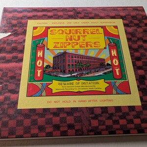 May include: A vintage Squirrel Nut Zippers album cover. The cover features a red and black checkered pattern with a yellow and red central design. The design includes the band's name, a building illustration, and the text "HOT".