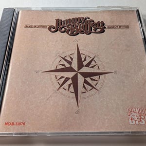 Pode incluir: Uma caixa de CD com o álbum "Changes in Latitudes, Changes in Attitudes" de Jimmy Buffett. A capa apresenta um design de rosa dos ventos em tons de castanho e o nome do artista em uma fonte estilizada. As palavras "Compact Price Disc" também são visíveis.
