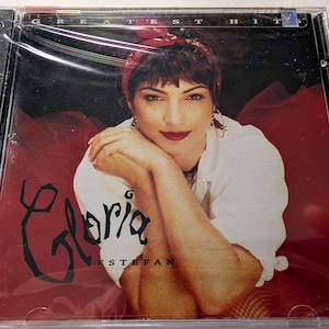 May include: Sealed CD of Gloria Estefan's "Greatest Hits" album. The cover features a portrait of Estefan in a white shirt and red headband, with the title "Gloria" in a stylized script. The background is a deep red.