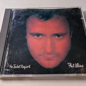May include: A CD case featuring a portrait of Phil Collins with a red-toned face against a black background. The album title, "No Jacket Required," is printed in white, along with the artist's name, "Phil Collins."