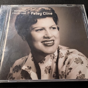 Puede incluir: Un CD de música en una caja de plástico transparente con un retrato en blanco y negro de Patsy Cline en la portada. El texto dice The Definitive Collection y Patsy Cline. El sujeto viste una camisa clara con un estampado floral oscuro.