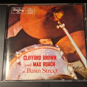 May include: A compact disc case featuring the album cover for Clifford Brown and Max Roach at Basin Street. The artwork displays a drummer behind a drum kit with a large cymbal, with the text Clifford Brown and Max Roach at Basin Street.