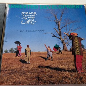 Puede incluir: Caja de CD del álbum "Arrested Development: 3 Years, 5 Months and 2 Days in the Life Of..." con un grupo de personas en un campo cubierto de hierba bajo un cielo azul. El título del álbum y el nombre del artista están impresos en la portada.