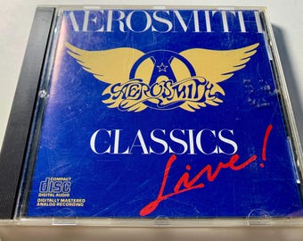 Puede incluir: CD "Classics Live!" de Aerosmith en una caja transparente. La portada del álbum es azul con el logo de la banda en dorado y las palabras "CLASSICS Live!" en blanco y rojo.