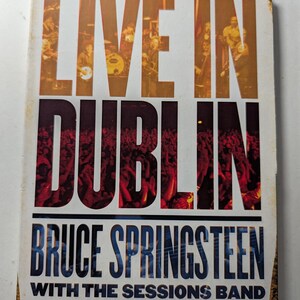 Puede incluir: Caja de DVD de "Live in Dublin" de Bruce Springsteen con The Sessions Band. La portada presenta el título en letras grandes de color naranja sobre una imagen de concierto, con "Dublin" en rojo y el nombre del artista en azul.