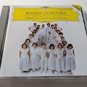 May include: A compact disc case featuring the album "Rossini Overtures" by the Orpheus Chamber Orchestra. The cover has a yellow banner with text and a group photo of the orchestra members in white outfits. The case is in a clear plastic jewel case.