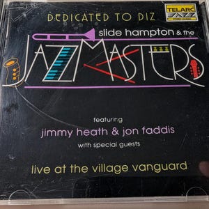 May include: A CD case with a black cover. The text reads "DEDICATED TO DIZ" and "slide hampton & the JAZZMASTERS." The cover features musical instrument graphics and the names "jimmy heath & jon faddis." The CD is labeled "live at the village vanguard."