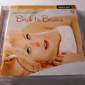 May include: A CD case featuring the album "Back to Basics" by Christina Aguilera. The cover art shows a close-up of a woman with blonde hair and red lipstick. The CD case is clear plastic, and the album title is in a stylized font.