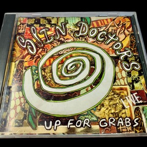 Pode incluir: Uma caixa de CD com o álbum "Up For Grabs" de Sin Doctors. A capa apresenta um design em espiral branco e verde, com o nome da banda em uma fonte estilizada. O fundo é uma colagem de cores.