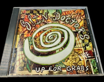 Puede incluir: Una caja de CD con el álbum "Up For Grabs" de Sin Doctors. La portada presenta un diseño en espiral blanco y verde, con el nombre de la banda en una fuente estilizada. El fondo es un collage de colores.