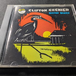 May include: A Clifton Chenier Bayou Blues compact disc in a clear plastic jewel case. The cover art features a black heron silhouette against a red and orange sunset, a yellow sun, and a green marsh landscape with a small cabin and blue text.