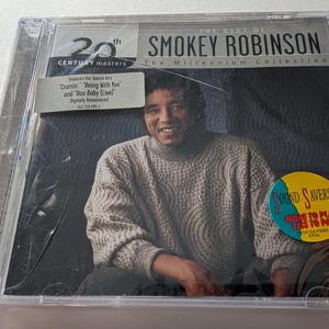 Puede incluir: Una caja de CD con el álbum "The Best Of" de Smokey Robinson. La portada muestra un retrato sonriente de Smokey Robinson con un suéter de color claro. La caja del CD tiene una etiqueta "20th Century Masters" y una pegatina "Sound Savers".