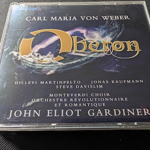 May include: A compact disc case featuring the opera Oberon by Carl Maria von Weber. The cover art displays a dark blue background with gold lettering, a large stylised O, and the names Hillevi Martinpelto, Jonas Kaufmann, Steve Davislim, Monteverdi Choir, Orchestre Révolutionnaire et Romantique, and John Eliot Gardiner.