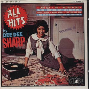 May include: A red and white starburst design with the text "All the Hits by Dee Dee Sharp" and "Volume 23" on a white background. A woman in a grey dress with a white collar and bow sits on a patterned carpet with a record player and a stack of records.
