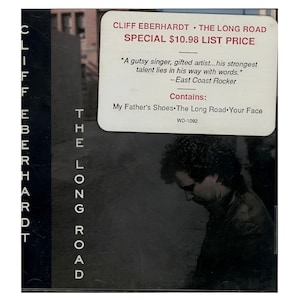 May include: CD case for Cliff Eberhardt's album, "The Long Road." The cover features the artist's name and album title in white text. A sticker displays the album's title, price, and a quote. The image includes a photo of the artist.