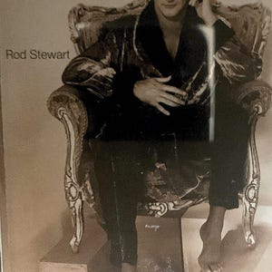 May include: A sepia-toned image featuring Rod Stewart seated in an ornate, gilded chair. He wears a dark velvet jacket and pants. The text "Rod Stewart" is visible in the upper left corner. He is barefoot and appears to be on a stage.