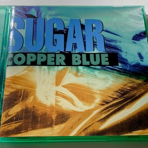 Puede incluir: Una caja de CD con el título del álbum "SUGAR COPPER BLUE" en letras azules grandes. El fondo tiene una mezcla de colores azul, cobre y blanco. La caja del CD es verde.