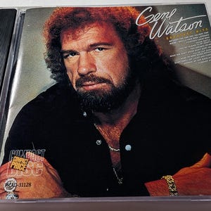 May include: A Gene Watson "Greatest Hits" CD album in a clear plastic case. The album cover features a portrait of Gene Watson with text that reads "Greatest Hits" and "MCA Gold Hits". The CD case has a "Compact Price" sticker.